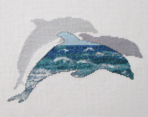 Cross stitch pattern of the silhouette of a dolphin filled with a scene of blue and turquoise ocean waves, with two plain silhouetted dolphins behind it. Straight view of stitched piece.