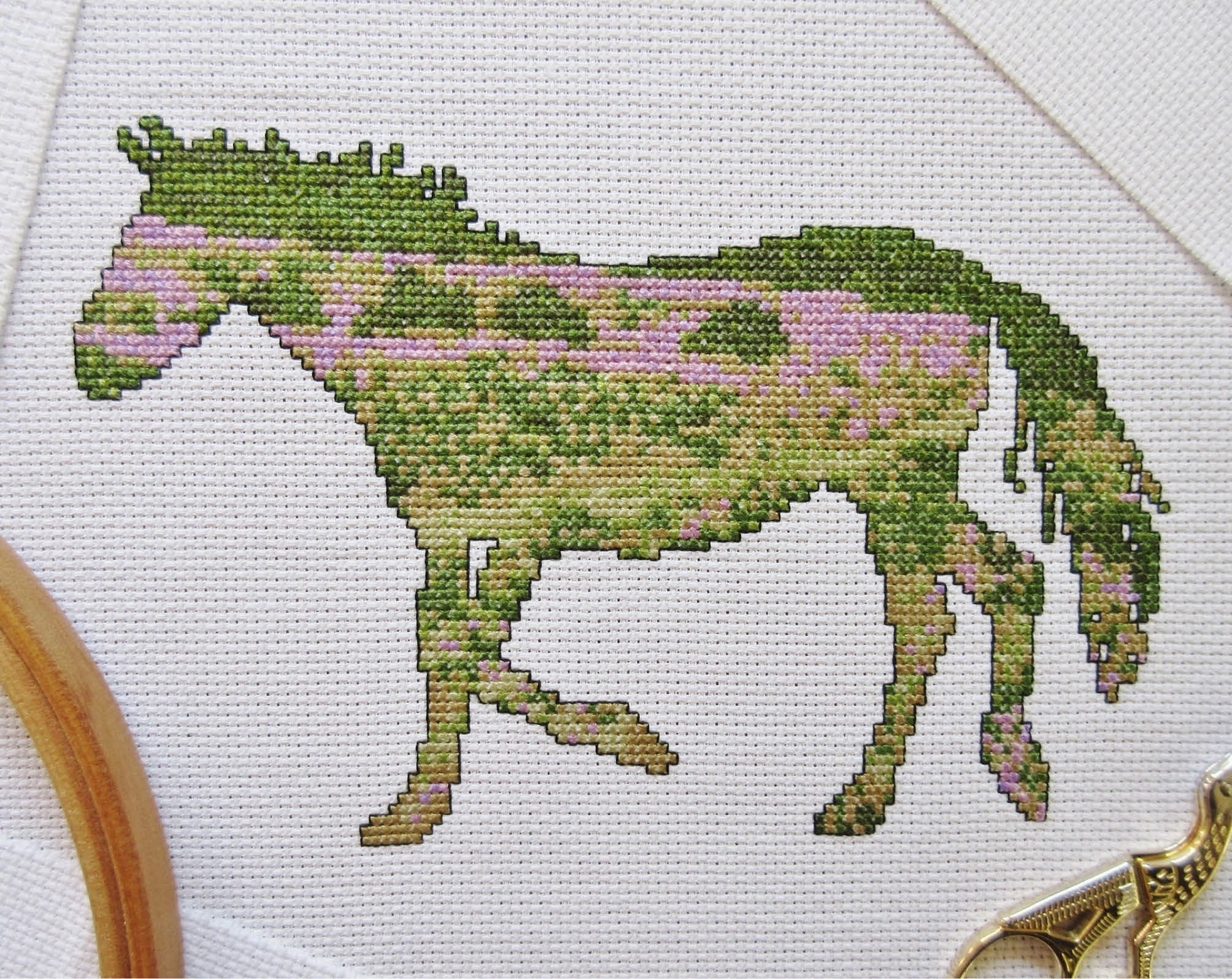 Cross stitch pattern of the silhouette of a pony with a view of the New Forest in southern England inside it. Stitched piece with hoop and sewing scissors.