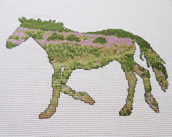 Cross stitch pattern of the silhouette of a pony with a view of the New Forest in southern England inside it. Angled view of stitched piece.