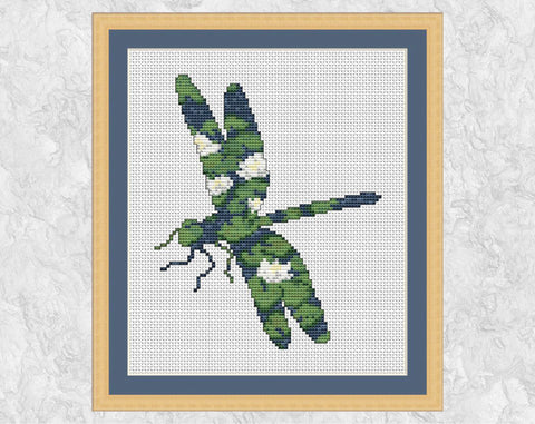 Cross stitch pattern of the silhouette of a dragonfly, filled with a peaceful scene of waterlilies on a pond. Shown with frame.