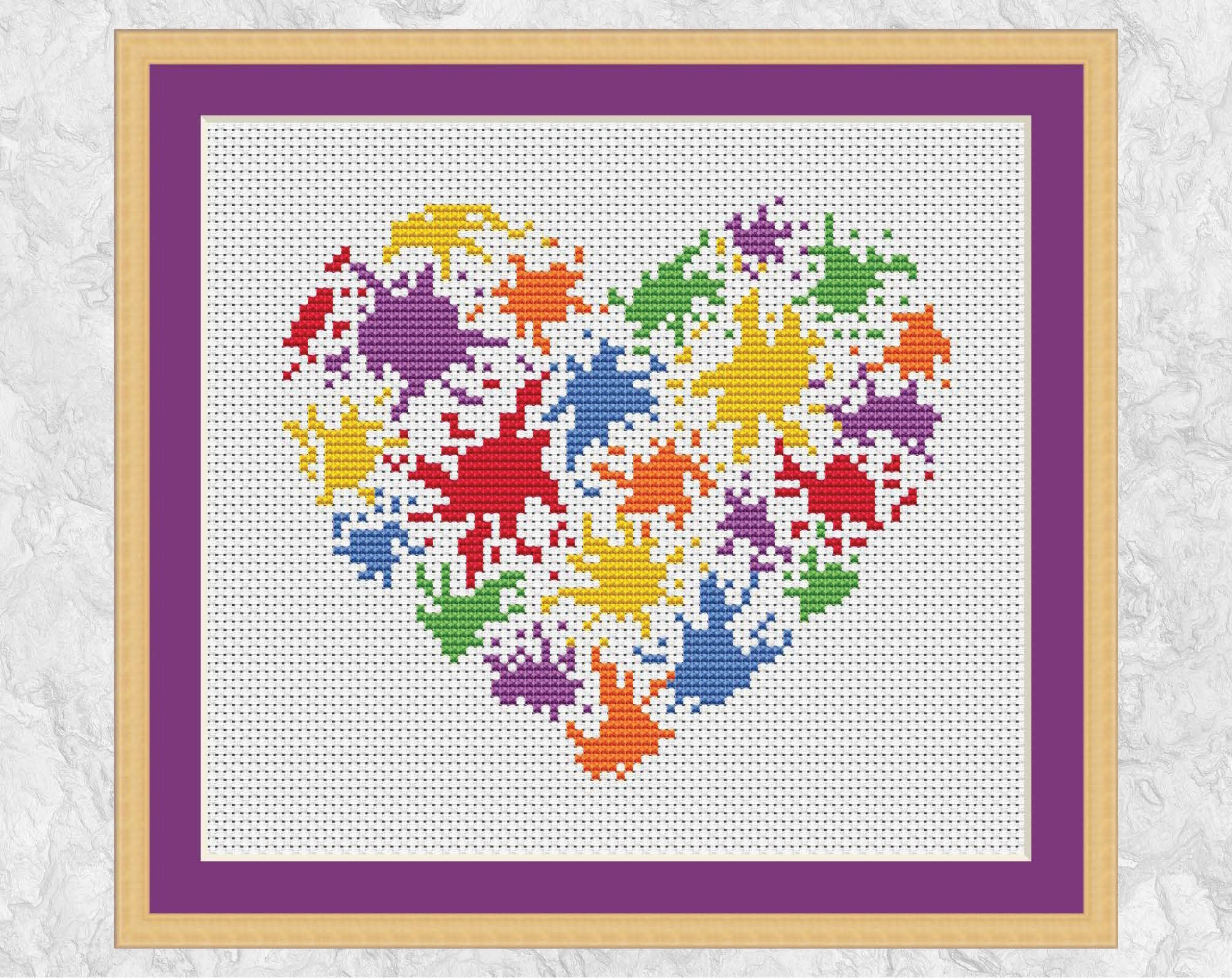 Bright cross stitch pattern of a splattered paint heart in rainbow colours. Shown with frame.