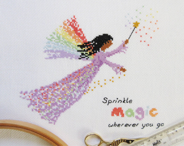 Rainbow Fairy cross stitch pattern - fairy in a purple dress with a wand and wings made of rainbows, and the text 'sprinkle magic wherever you go'. Stitched piece with props.