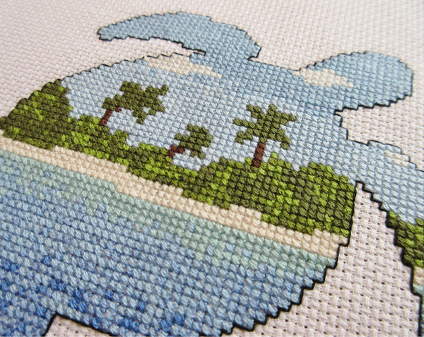 Cross stitch pattern of the silhouette of a turtle filled with a view of a desert island beach, blue sea and sky. Close up angled view of stitched piece.