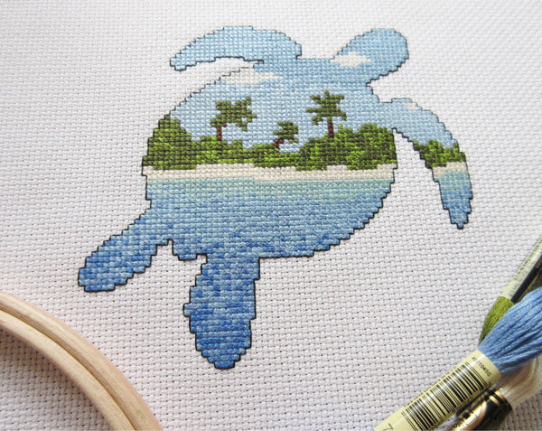 Cross stitch pattern of the silhouette of a turtle filled with a view of a desert island beach, blue sea and sky. Angled view of stitched piece with props.