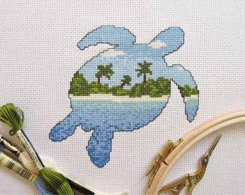 Cross stitch pattern of the silhouette of a turtle filled with a view of a desert island beach, blue sea and sky. Stitched piece with props.