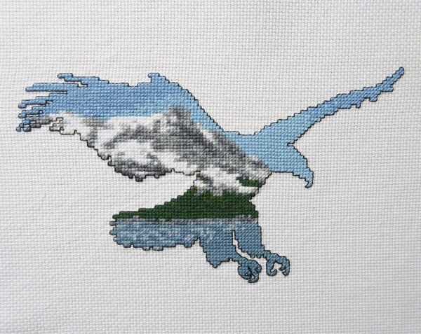 Cross stitch pattern of the silhouette of a bald eagle filled with a scene of snow covered mountains, forest and a lake. Stitched piece.