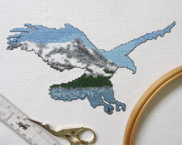 Cross stitch pattern of the silhouette of a bald eagle filled with a scene of snow covered mountains, forest and a lake. Stitched piece with props.