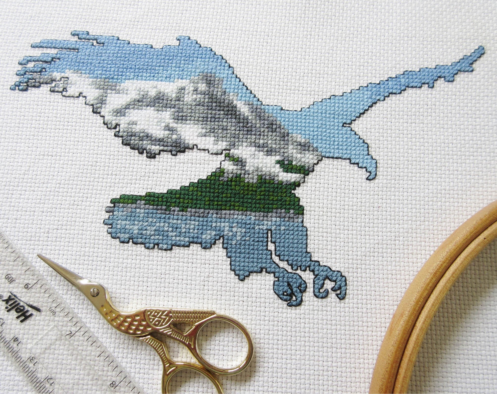 Cross stitch pattern of the silhouette of a bald eagle filled with a scene of snow covered mountains, forest and a lake. Stitched piece with props.