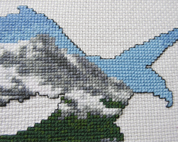Cross stitch pattern of the silhouette of a bald eagle filled with a scene of snow covered mountains, forest and a lake. Close up view of stitched piece.