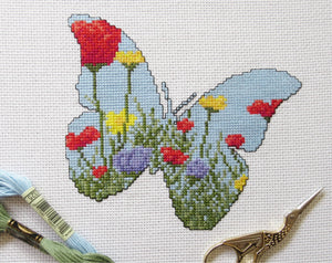 Cross stitch pattern of the silhouette of a butterfly, filled with a scene of a wildflower meadow of grasses, poppies, dandelions and cornflowers. Stitched piece with prop.