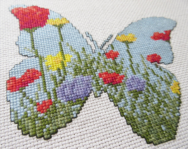 Cross stitch pattern of the silhouette of a butterfly, filled with a scene of a wildflower meadow of grasses, poppies, dandelions and cornflowers. Angled view of stitched piece.