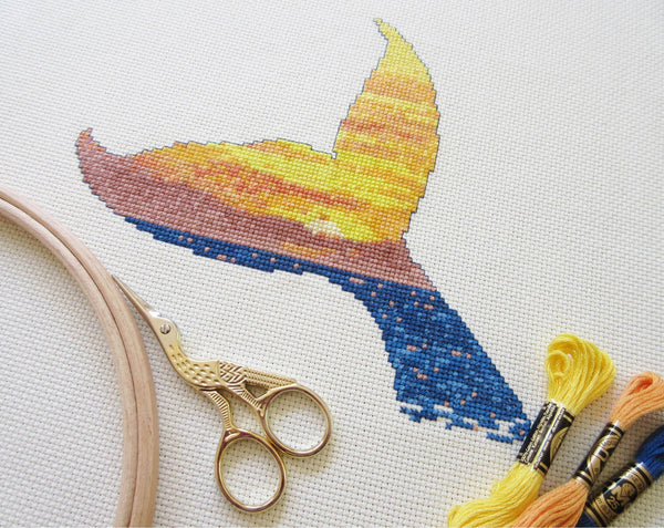 Cross stitch pattern of the silhouette of a whale's tail filled with a scene of a sunset over the ocean. Angled view of stitched piece with sewing scissors, hoop and threads.