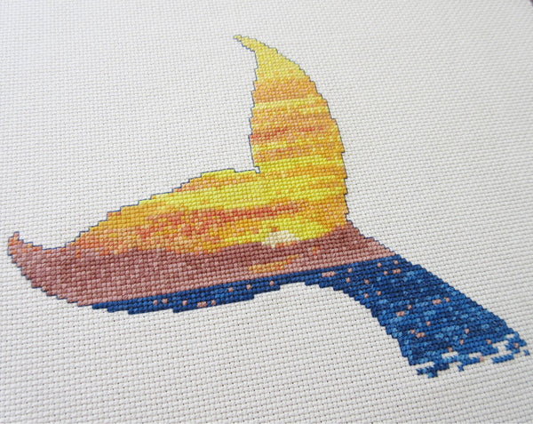 Cross stitch pattern of the silhouette of a whale's tail filled with a scene of a sunset over the ocean. Angled view of stitched piece.
