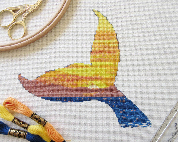 Cross stitch pattern of the silhouette of a whale's tail filled with a scene of a sunset over the ocean. Stitched piece with props.