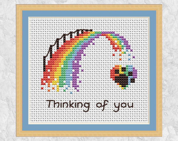 Cross stitch pattern PDF of a rainbow bridge ending at a paw print on a heart, with the words "Thinking of you". Shown with frame.