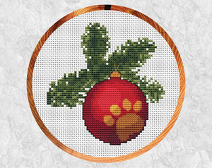 Cross stitch pattern of a bauble engraved with a paw print hanging from a Christmas tree branch. Shown in hoop.