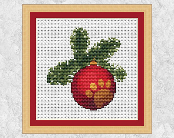 Cross stitch pattern of a bauble engraved with a paw print hanging from a Christmas tree branch. Shown with frame.