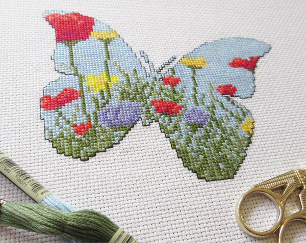 Cross stitch pattern of the silhouette of a butterfly, filled with a scene of a wildflower meadow of grasses, poppies, dandelions and cornflowers. Angled view of stitched piece with props.