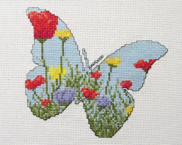 Cross stitch pattern of the silhouette of a butterfly, filled with a scene of a wildflower meadow of grasses, poppies, dandelions and cornflowers. Straight view of stitched piece.