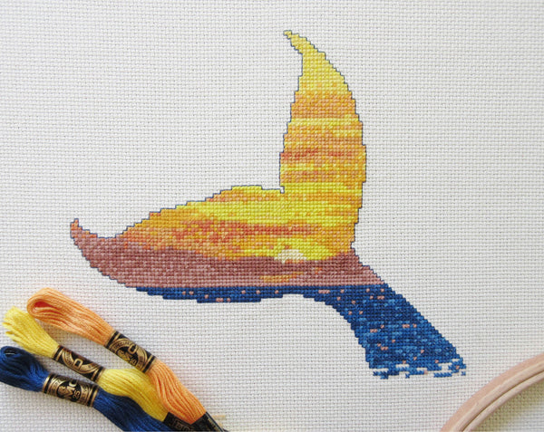 Cross stitch pattern of the silhouette of a whale's tail filled with a scene of a sunset over the ocean. Stitched piece with DMC threads and embroidery hoop.