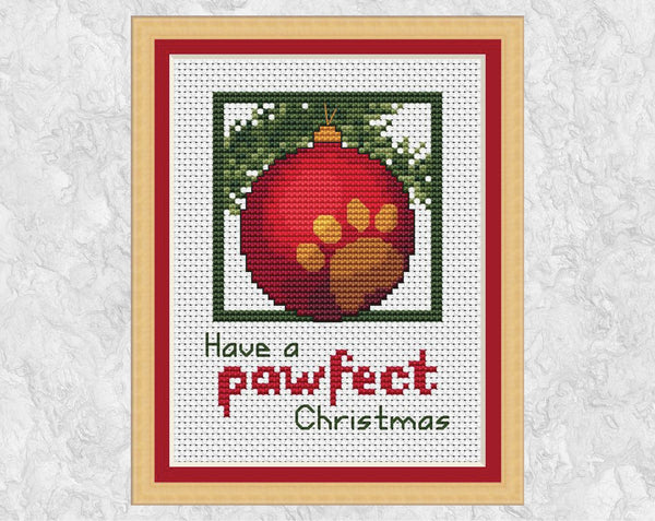 Cross stitch pattern of a red bauble with a gold paw print on it, hanging from a branch with the words 'Have a pawfect Christmas'. Shown with frame.