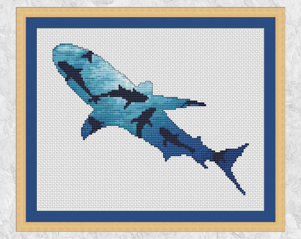 Cross stitch pattern of the silhouette of a shark, filled with a scene looking upwards through a shoal of sharks, with sunlight illuminating the water from above. Shown with frame.