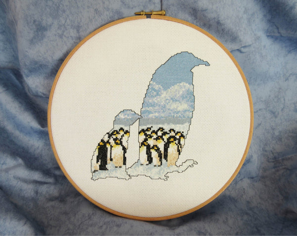 Cross stitch pattern of the silhouette of a penguin and chick, filled with an Antarctic scene of penguins huddled together on the snow, with mountains in the distance below a clear blue sky. Stitched piece in hoop.
