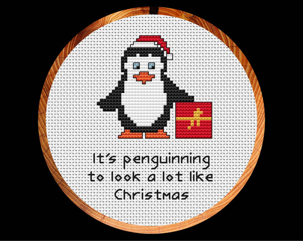 'It's Penguinning to Look a Lot Like Christmas' cross stitch pattern with penguin wearing Santa hat and holding present. Shown in hoop.