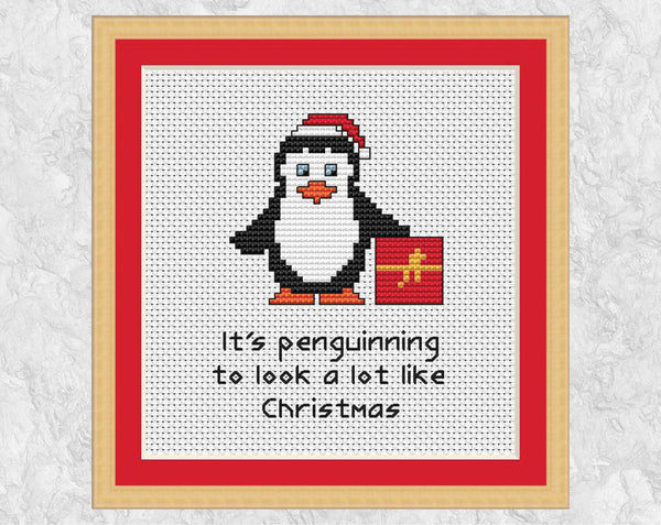 'It's Penguinning to Look a Lot Like Christmas' cross stitch pattern with penguin wearing Santa hat and holding present. Shown in frame.