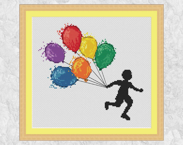 Cross stitch pattern of a boy running with watercolour effect balloons in rainbow colours. Shown with frame.