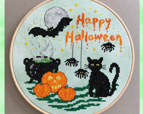 'A Halloween Night' cross stitch pattern - featuring a bat, moon, cauldron, pumpkins, black cat, spiders and the words 'Happy Halloween'. Close up photo.