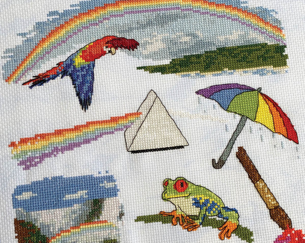 Colours of the Rainbow cross stitch pattern - rainbow, parrot, prism, umbrella, waterfall, frog and paints and paintbrush. Angled view of stitching.